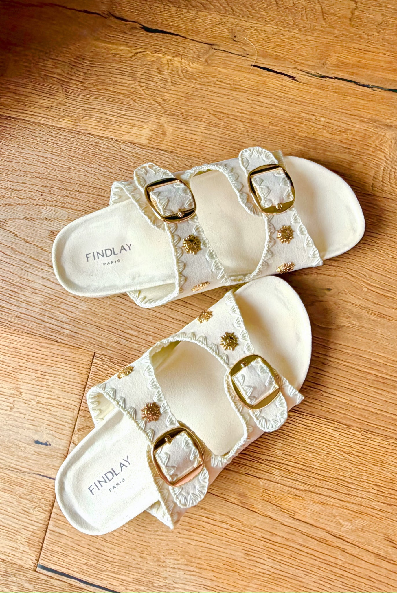 Golden Shore Slide Sandals von Findlay Paris in Ivory mit goldenen Schnallen auf einem Holzboden.