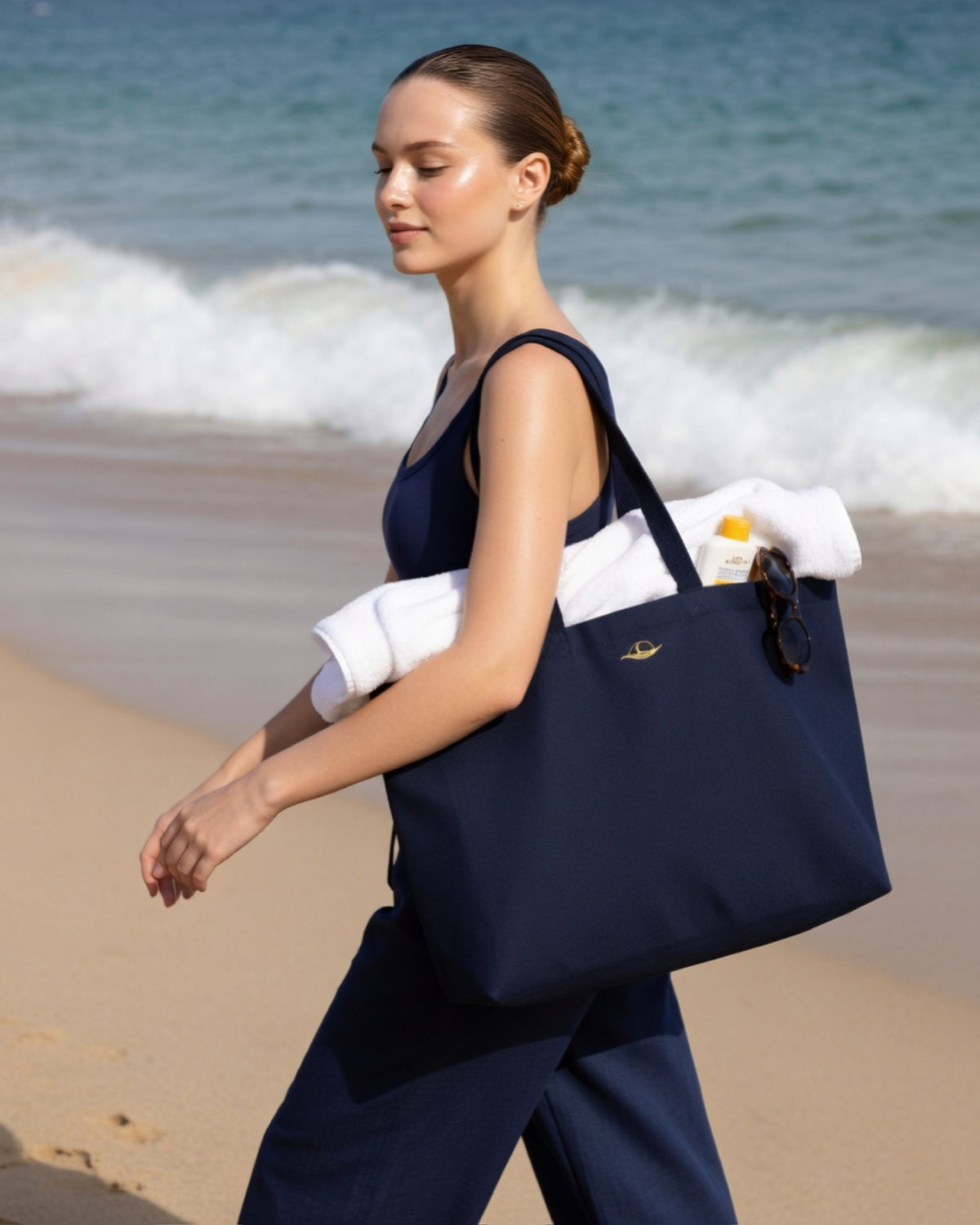 Frau am Strand mit der dunkelblauer Strandtasche, handgepäckt mit Handtüchern, lässig und elegant, bereit für den Sommer.