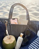 Gestreifte Strandtasche mit Handy, Sonnenbrille, Kokosnuss und Sonnencreme am Strand.