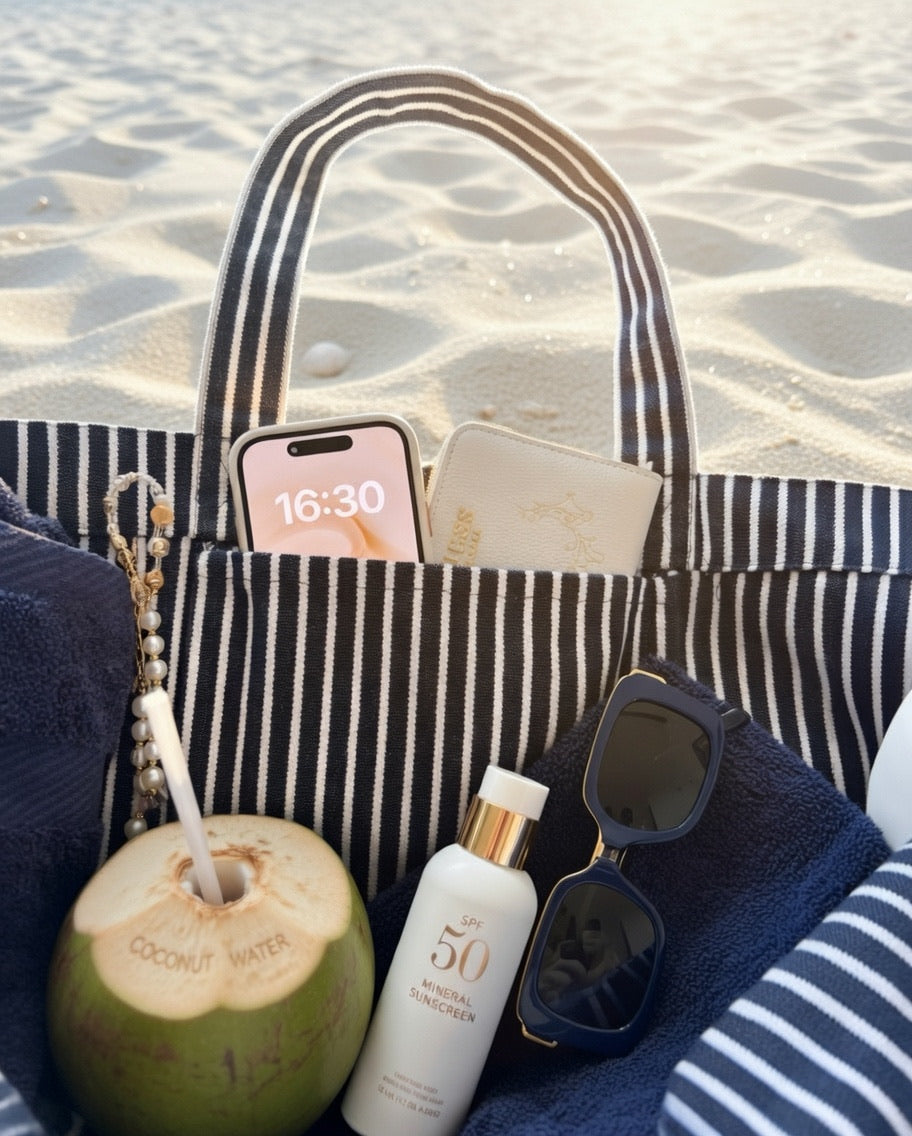 Gestreifte Strandtasche mit Handy, Sonnenbrille, Kokosnuss und Sonnencreme am Strand.